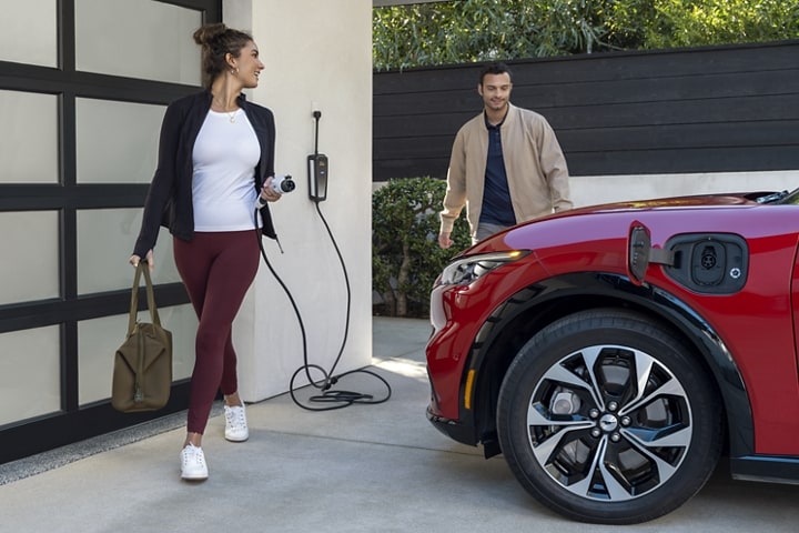 Woman holding a charger and looking back at a man who is gazing adoringly at the front end of the 2023 Ford Mustang Mach-E®