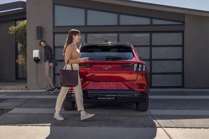 Woman walking around the rear of a 2023 Ford Mustang Mach-E® parked in the driveway as a man unplugs the vehicle