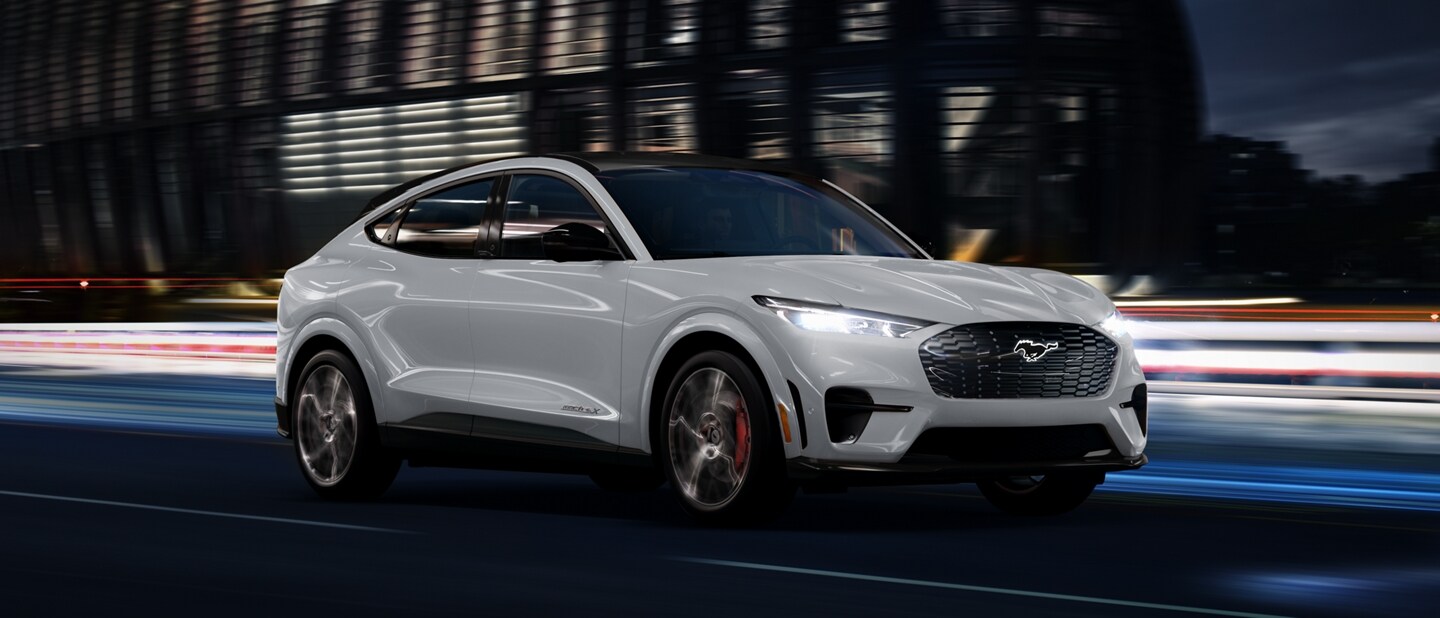 Space White Metallic 2023 Ford Mustang Mach-E® SUV being driven on a highway past a stadium at night