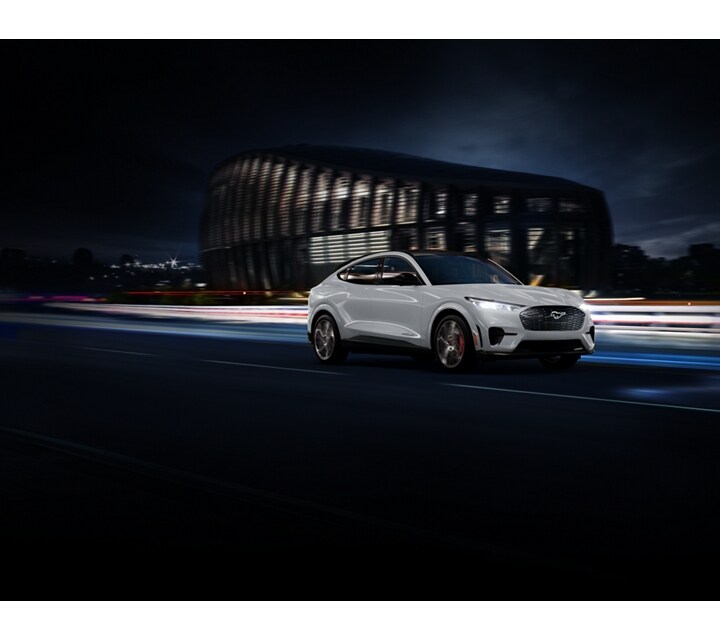 Space White Metallic 2023 Ford Mustang Mach-E® SUV being driven on a highway past a stadium at night