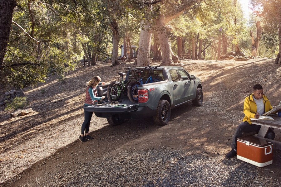 Woman at a campsite plugs a bike into the 400-watt 110V Outlet in the bed of a 2023 Ford Maverick® XLT truck