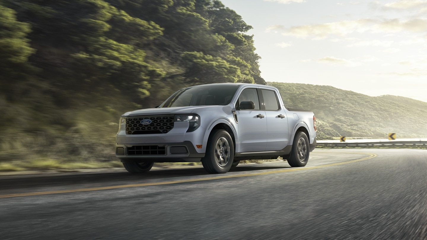 A white 2025 Ford Maverick® pickup driving on a winding highway near a lake in the forest