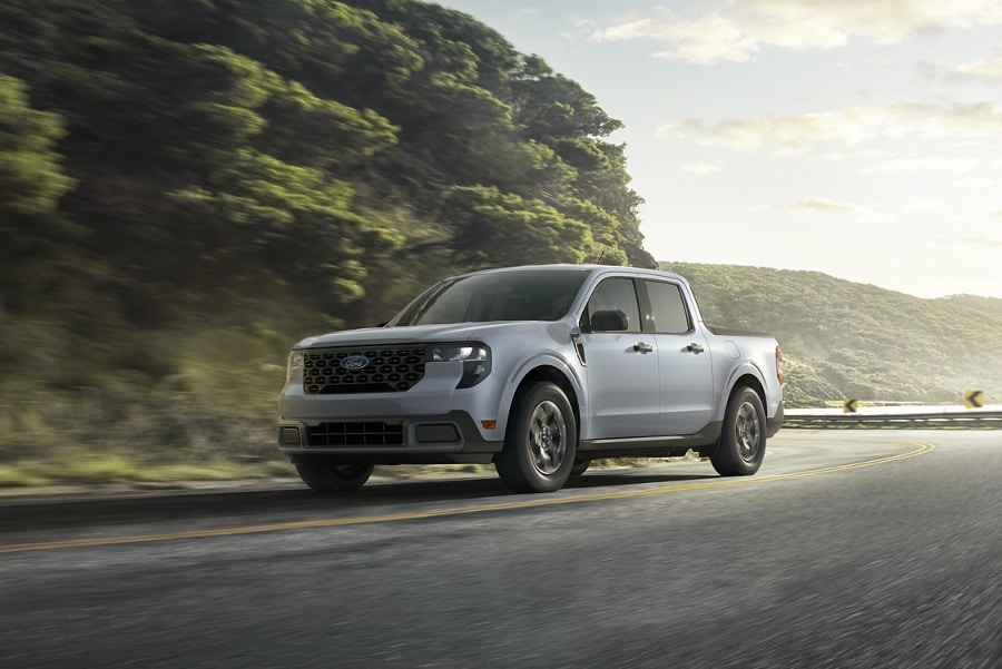 A white 2025 Ford Maverick® truck driving along a forest highway near a lake at sunset