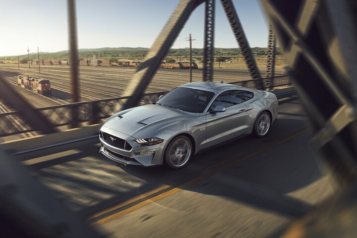 A 2023 Ford Mustang® GT model in Iconic Silver Metallic being driven across a metal bridge over a train yard
