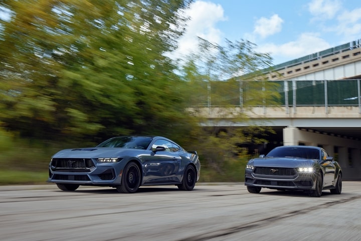 2024 Ford Mustang® GT coupe in Vapor Blue Metallic and an EcoBoost® model in Carbonized Grey Metallic being driven on a road