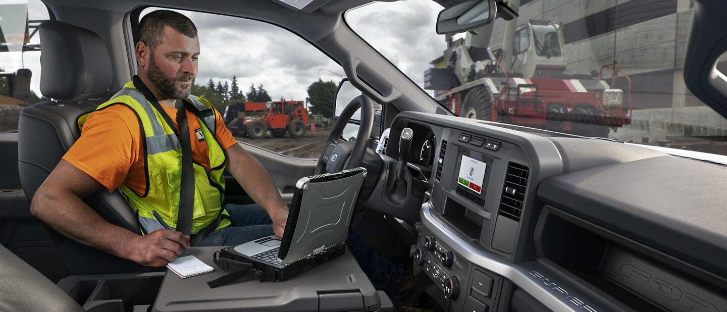 Man working on laptop inside a 2023 Ford Super Duty® F-250 XL with construction equipment in background
