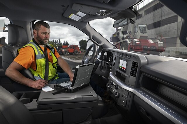 Man working on laptop inside a 2023 Ford Super Duty® F-250 XL with construction equipment in background