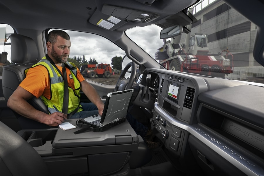 Man working on laptop inside a 2023 Ford Super Duty® F-250 XL pickup with a work site in background