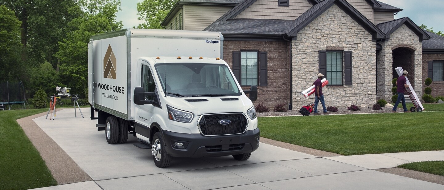 A 2023 Ford Transit® Chassis Cab parked in a driveway with people working on a house in the background