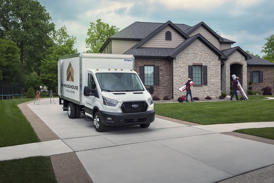 A 2023 Ford Transit® Chassis Cab parked in a driveway with people working on a house in the background
