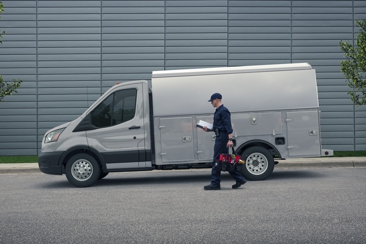 A mechanic’s 2023 Ford Transit® Chassis Cab upfit in front of large building with worker walking up to van