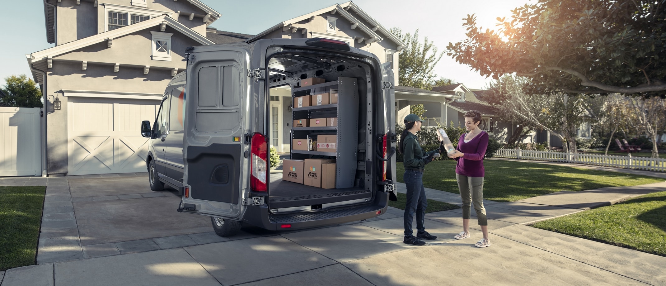 A 2024 Ford Transit® van parked in a driveway with rear doors open as a delivery person drops off a package