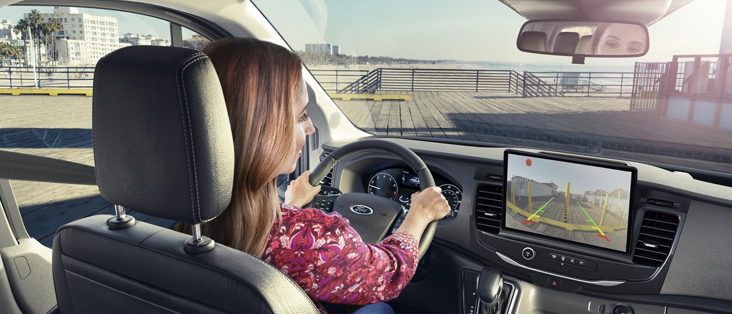 A woman driving a 2024 Ford Transit® van looking at her touchscreen seeing behind the van