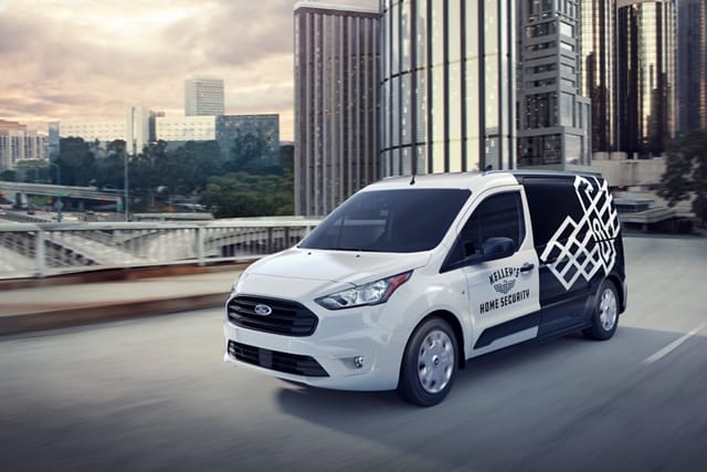 2023 Frozen White Transit Connect Cargo Van with aftermarket business graphics being driven on a city road