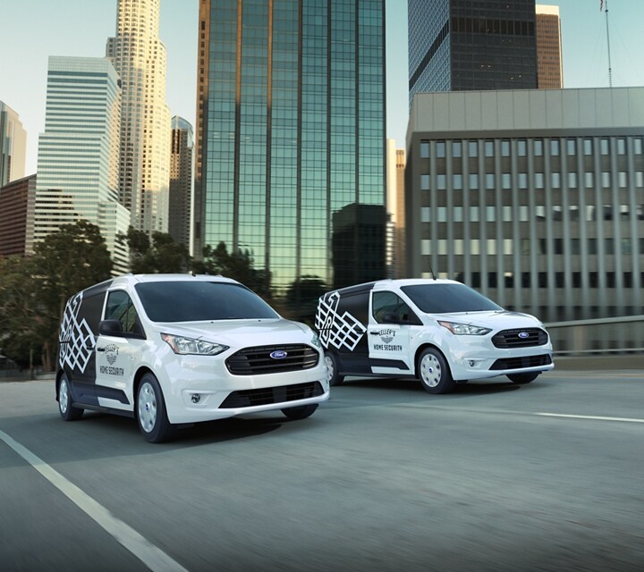 Two 2023 Ford Transit Connect Cargo Vans with eye-catching business graphics being driven on a city street